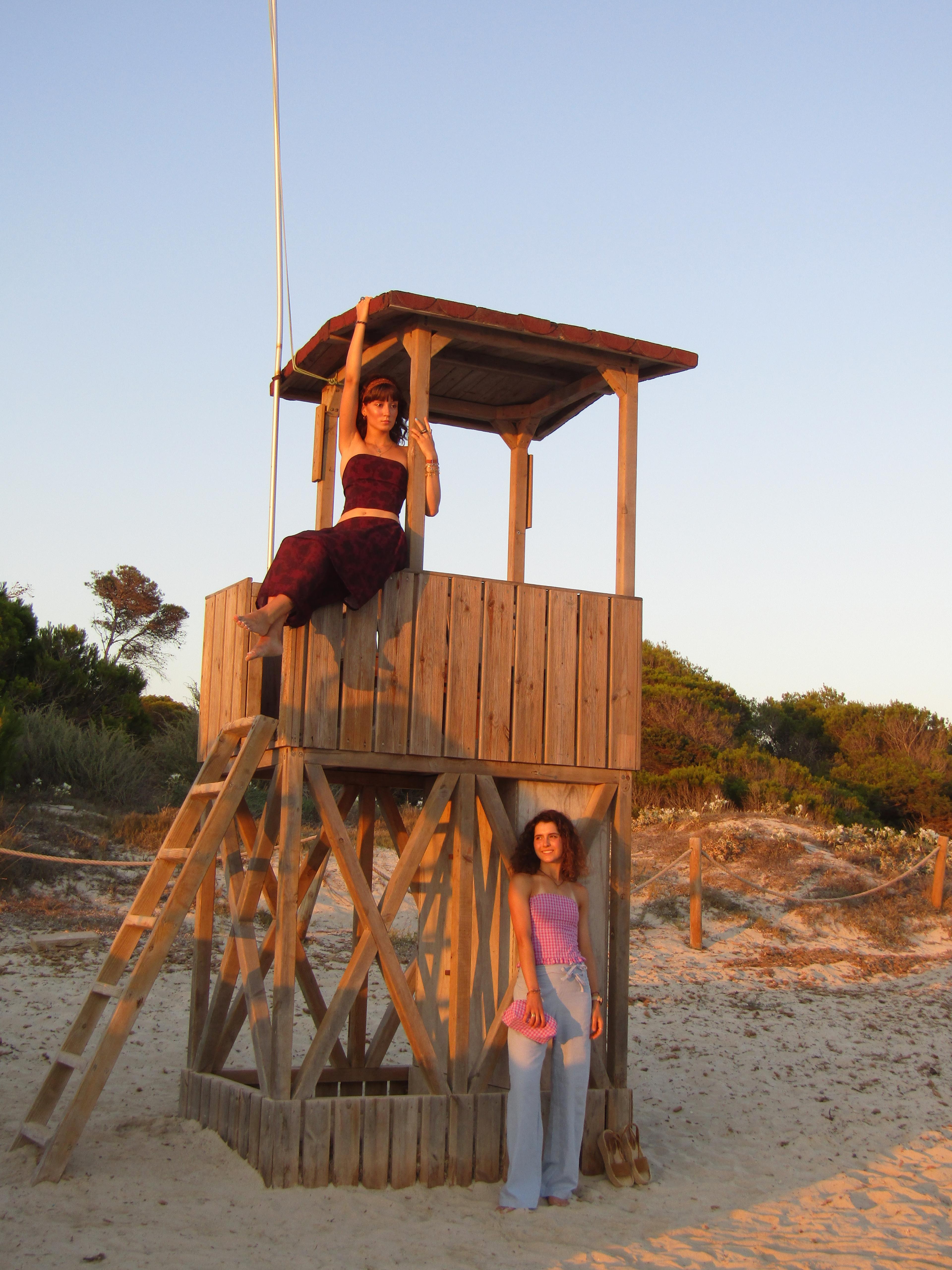 Foto en la playa de dos chicas, una subida encima de una torre de socorrista y la otra apoyada